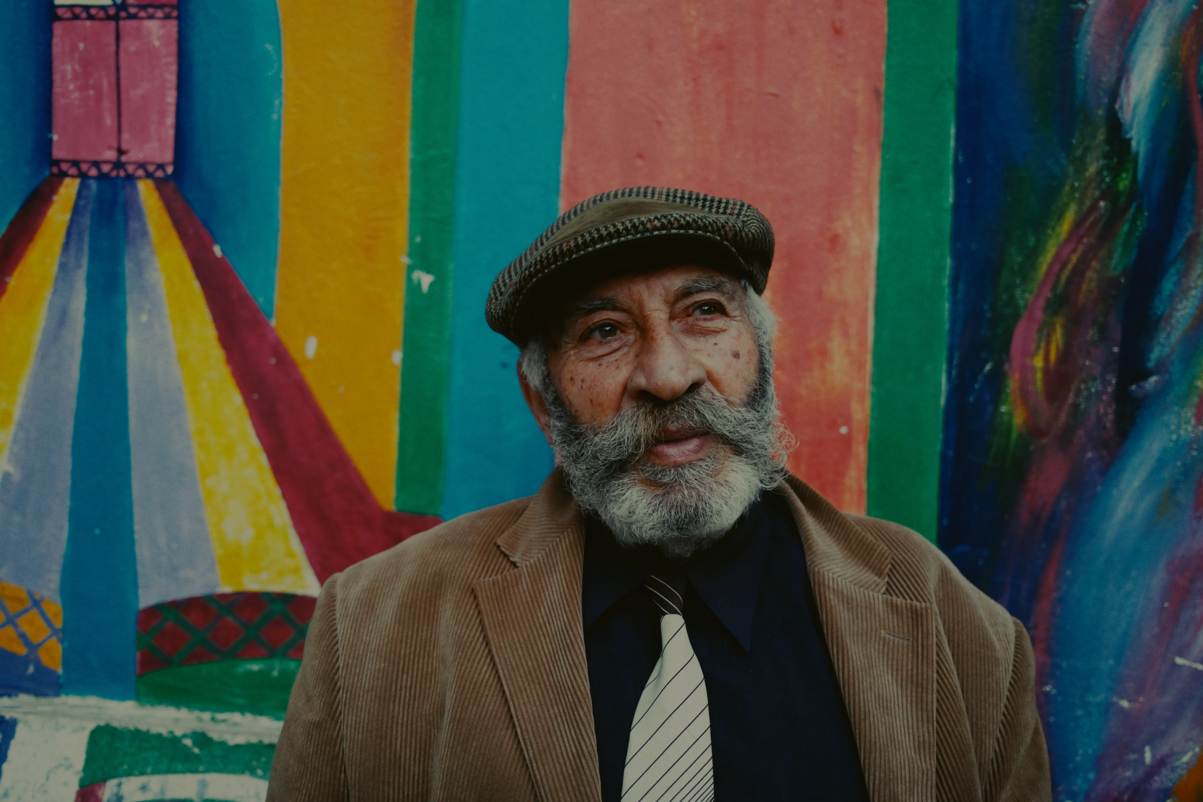 A man in a flat cap and corduroy jacket in front of a brightly painted wall, developed on Portra 400