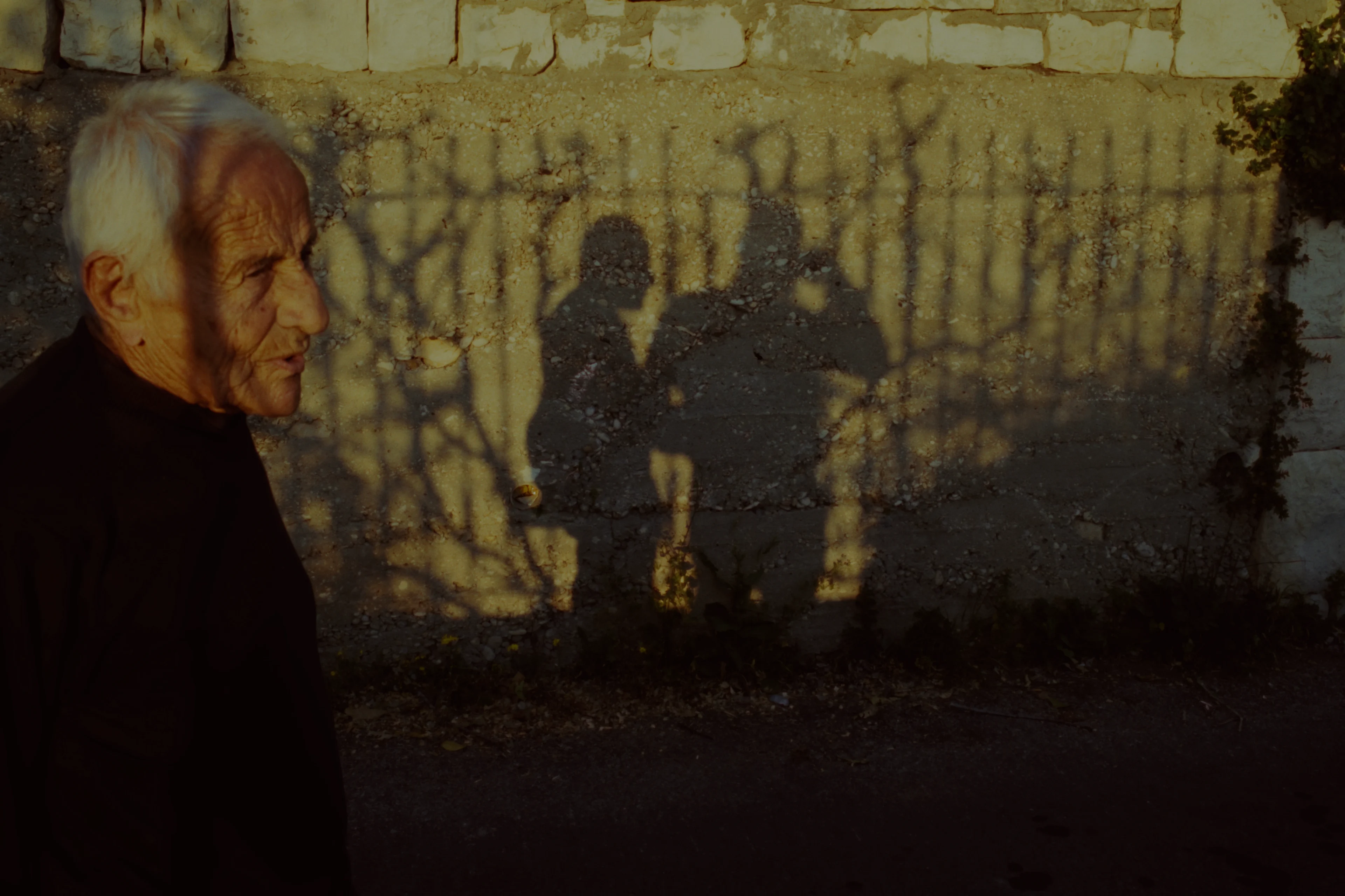 An older man walking past a sunlit wall carrying the shadows of two people and a fence, developed on Vision3 50D